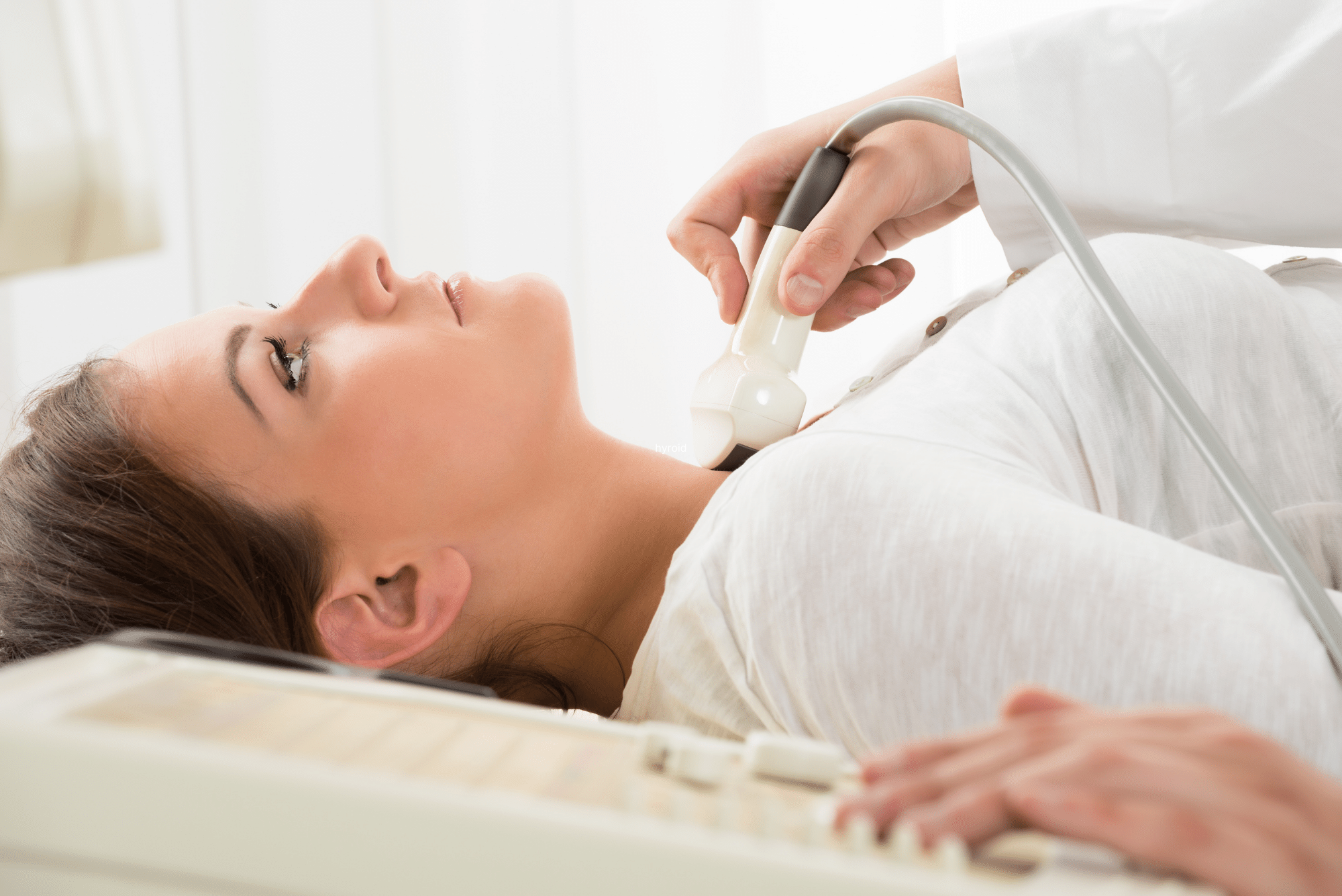 A woman in white reclines while a doctor uses ultrasound to check her thyroid.