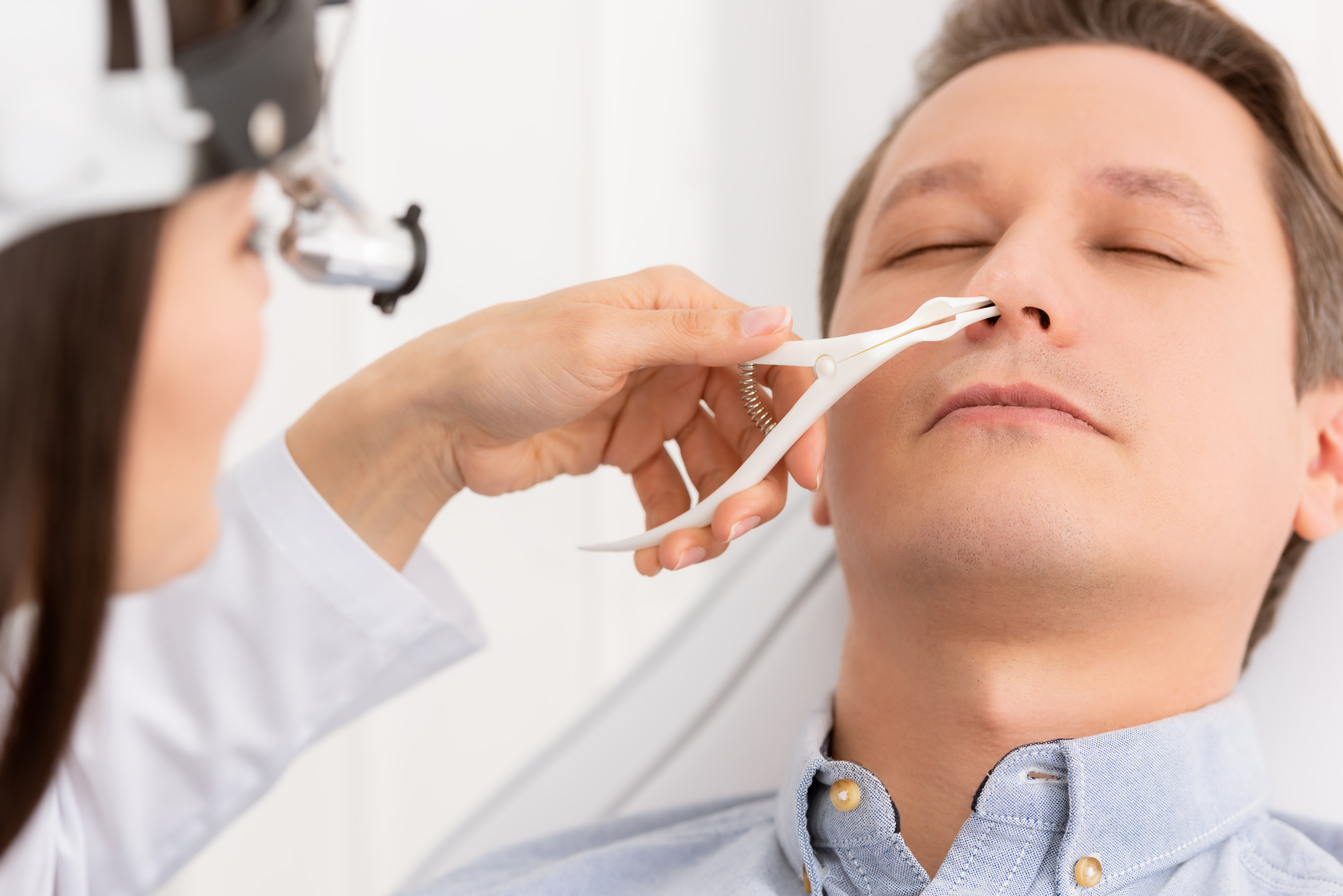 A doctor uses a nasal speculum to see into a reclining man's nostril.