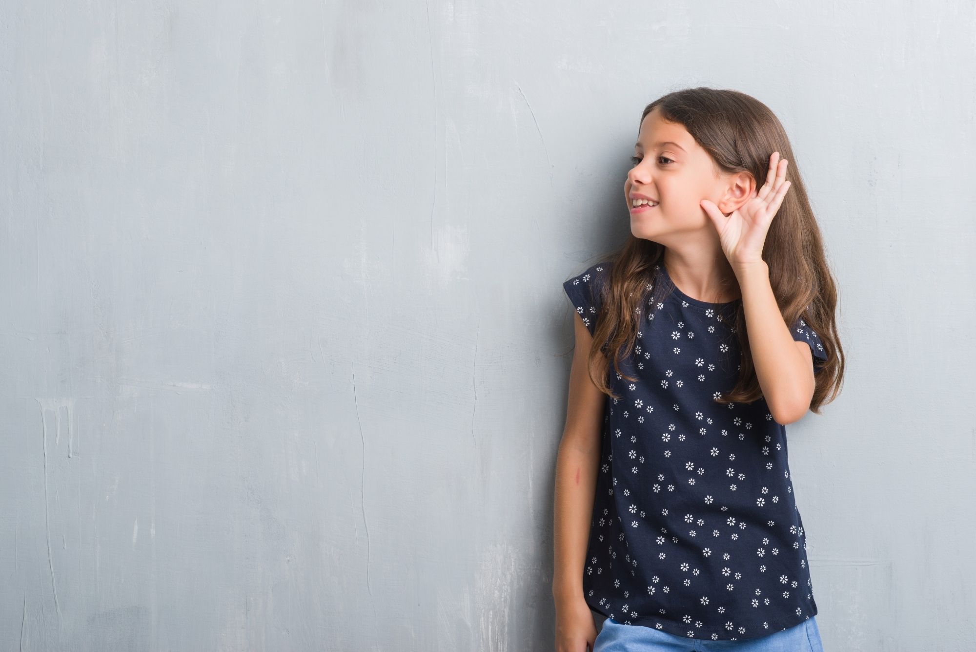 A little girl in a blue shirt against a concrete wall playfully cups her ear after getting tympanostomy tubes.