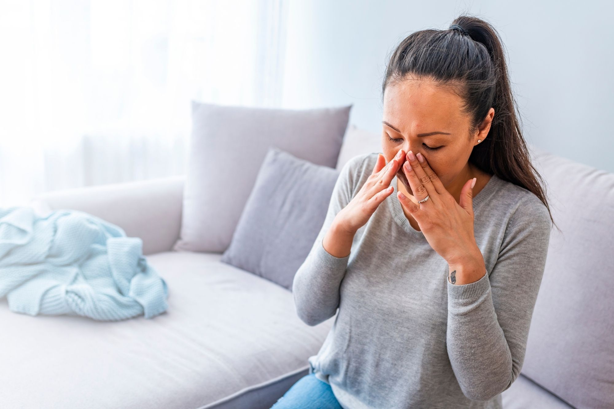 A woman sits on a grey couch, blowing her nose after taking the best antibiotic for a sinus infection.