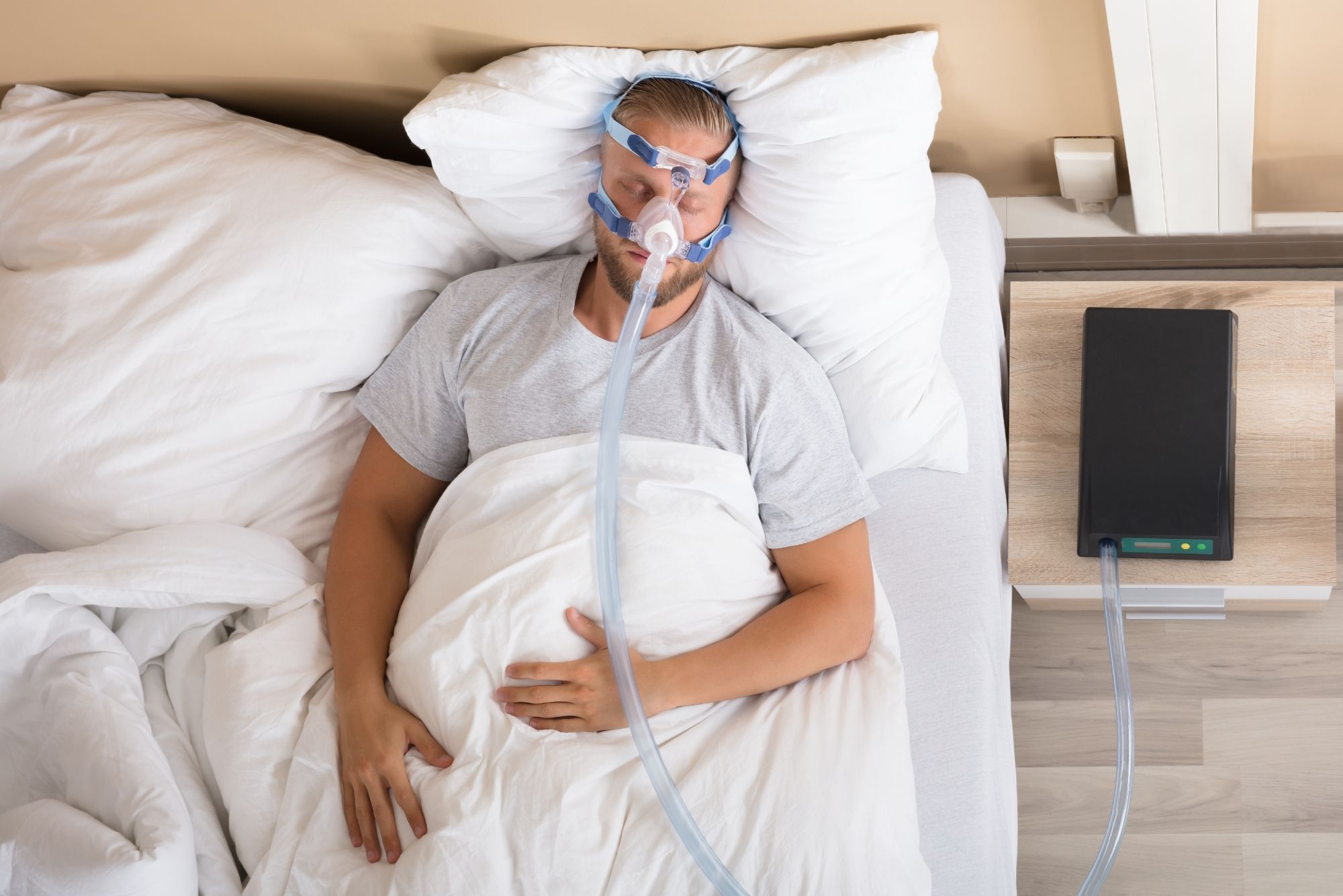 A man lies in a white bed wearing a sleep apnea machine.