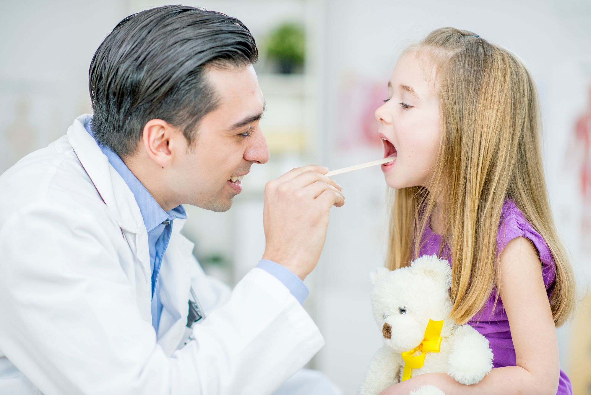 A little girl holding a stuffed bear gets her throat looked at by a pediatric otolaryngologist.