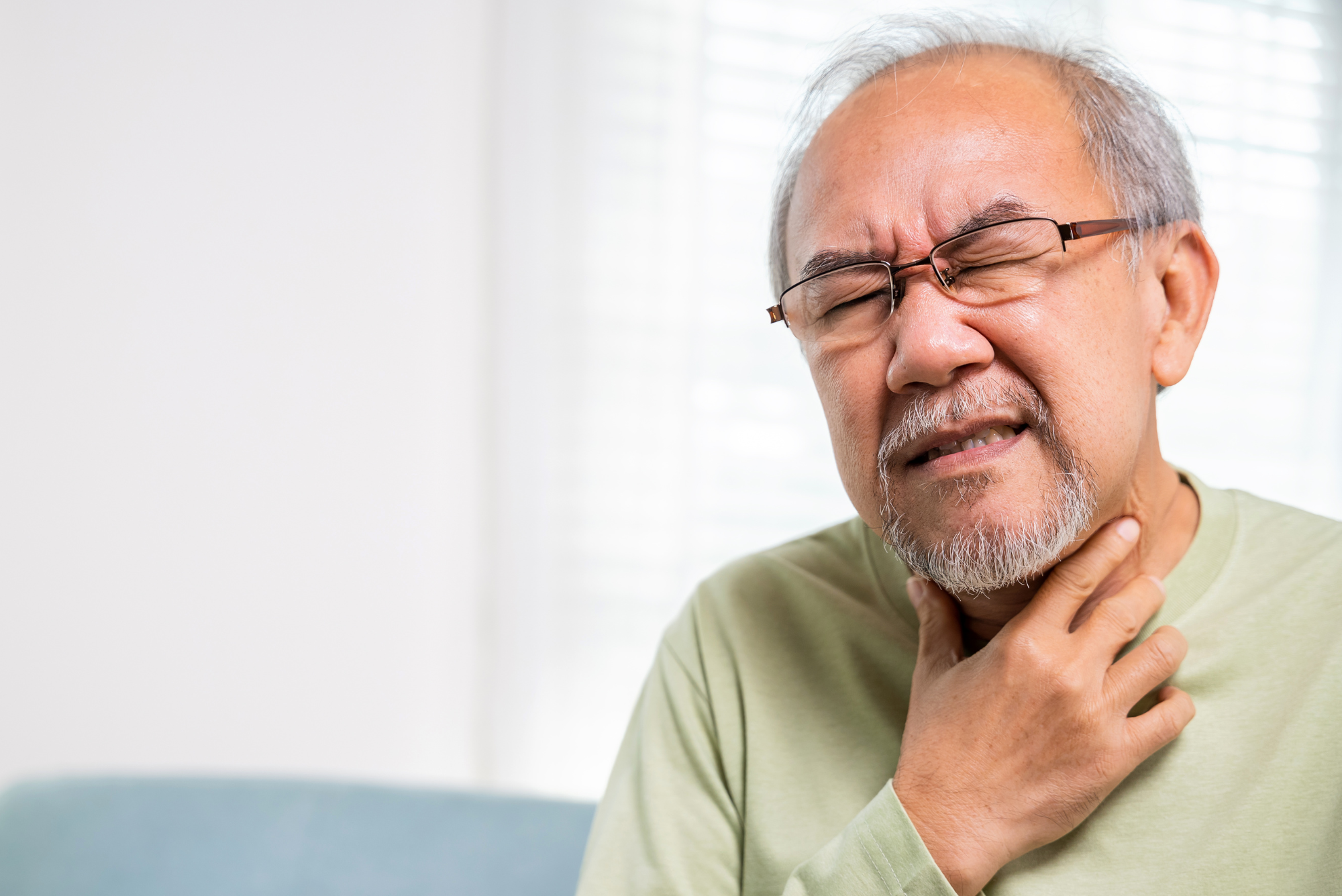 An older man in a green shirt holds his throat in pain before getting a vocal cord stripping procedure.