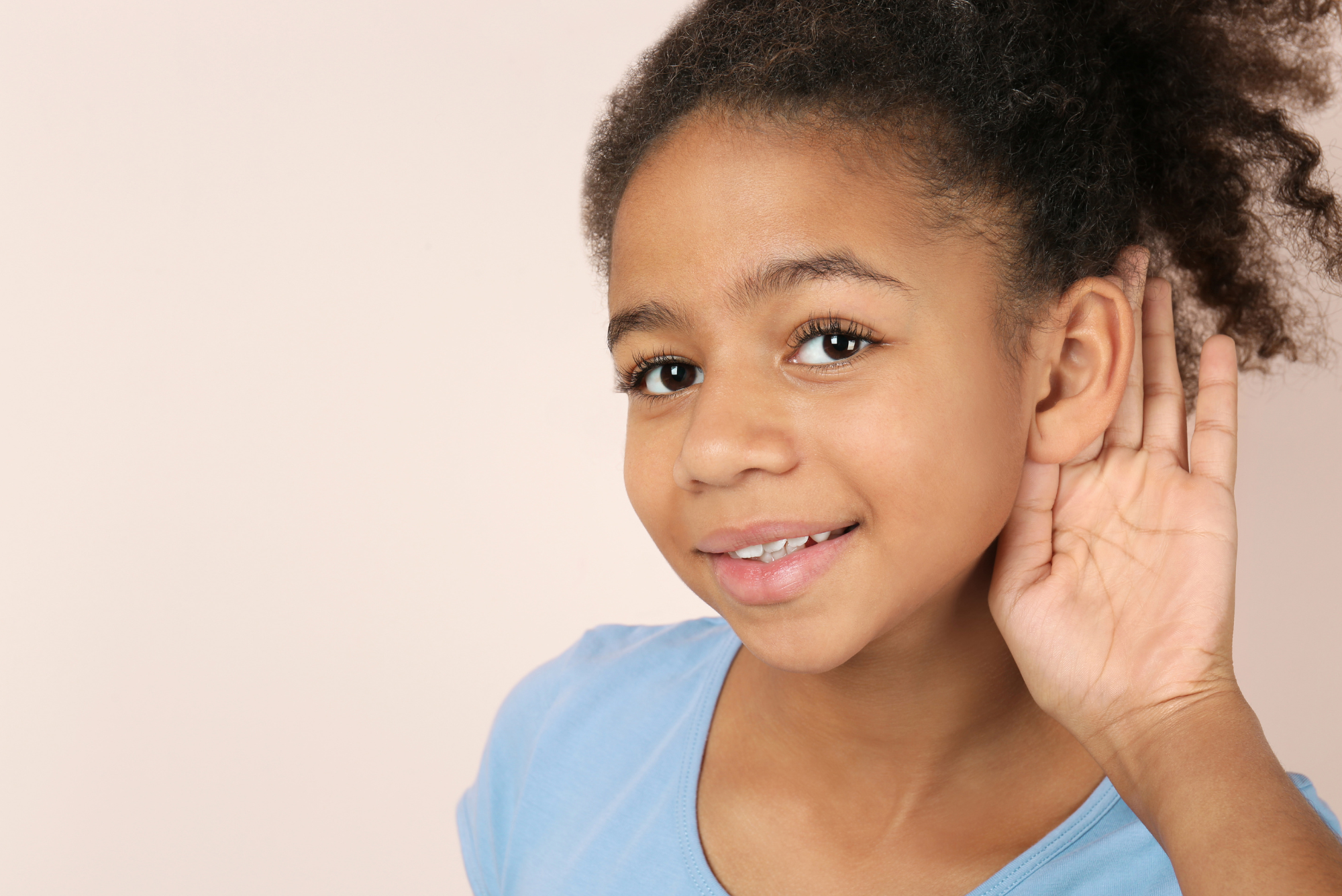 A young girl in a blue shirt holds her hand behind her ear in a listening position, representing ear tubes and adenoid removal.